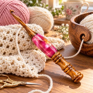 May include: A crochet hook featuring a polished wooden handle with a central section of marbled fuchsia and gold resin. The hook has a tapered wooden tip and a contoured grip with multiple ridges, resting on a cream-colored crocheted fabric.