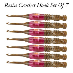 May include: Set of seven crochet hooks made of resin and wood. The hooks have a pink and brown color scheme. The hooks are arranged in a row, with the largest hook at the top and the smallest hook at the bottom.