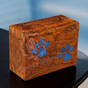 Könnte beinhalten: Eine hölzerne Tierurne mit blauen Pfotenabdrücken, die in das Holz eingelegt sind. Die Urne hat eine natürliche Holzoptik und wird von Woodco Handcrafts hergestellt.