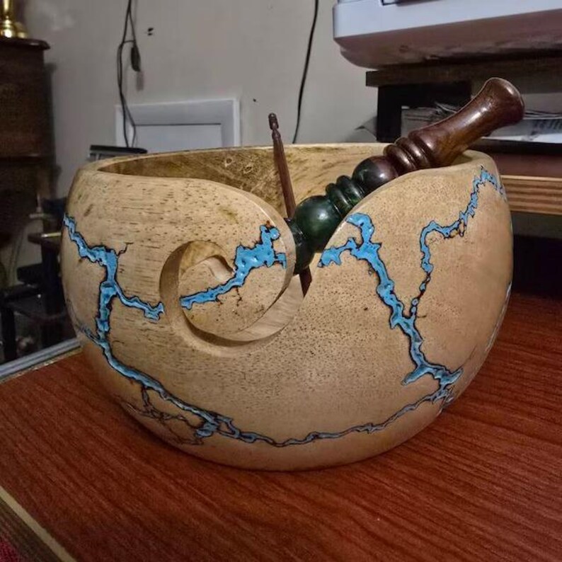 K&ouml;nnte beinhalten: Eine Garnschale aus hellem Holz mit einem spiralf&ouml;rmigen Ausschnitt und komplizierten, blitzartigen Rissen, die mit leuchtend blauem Harz gef&uuml;llt sind. Eine dunkelbraune H&auml;kelnadel aus Holz liegt in der Schale und ragt aus der &Ouml;ffnung.