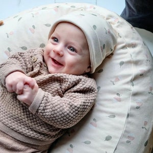 Puede incluir: Un bebé sonriente que lleva un suéter de punto marrón y un gorro blanco, acostado sobre una almohada de color crema con un patrón floral.