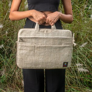 May include: A woman is holding a light beige hemp laptop bag with a zipper and a small logo on the bottom right corner. The bag is made of natural hemp fabric and has a woven texture.