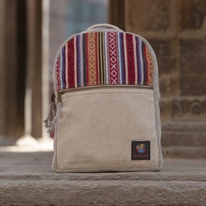 Op de afbeelding: Een beige canvas rugzak met een kleurrijk geweven patroon op de bovenklep. De rugzak heeft een ritsvak aan de voorkant en een klein logo-patch met de tekst "Buddhist Co."