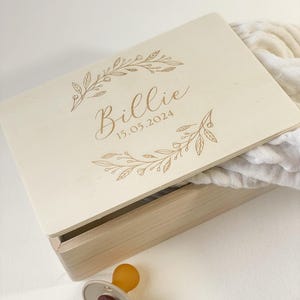 Puede incluir: Una caja de madera clara con el nombre "Billie" y la fecha "15.05.2024" grabados en la tapa, enmarcados por diseños florales. Un chupete con tetina marrón y anillo gris en primer plano. La caja está parcialmente abierta.