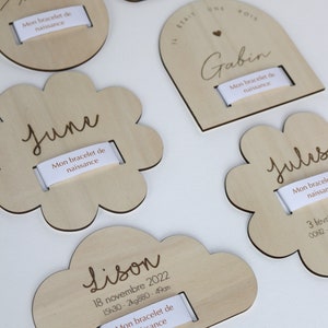 Puede incluir: Cinco placas de madera con diferentes formas y dise&ntilde;os, cada una con una ranura para un brazalete. Las placas est&aacute;n grabadas con los nombres "Ambre", "Augustin", "June", "Gabin" y "Jules", junto con las fechas y horas de nacimiento. Las placas est&aacute;n hechas de madera clara y tienen un acabado natural.