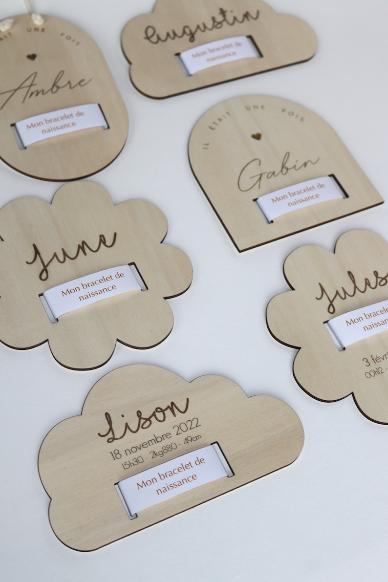 Puede incluir: Cinco placas de madera con diferentes formas y dise&ntilde;os, cada una con una ranura para un brazalete. Las placas est&aacute;n grabadas con los nombres "Ambre", "Augustin", "June", "Gabin" y "Jules", junto con las fechas y horas de nacimiento. Las placas est&aacute;n hechas de madera clara y tienen un acabado natural.