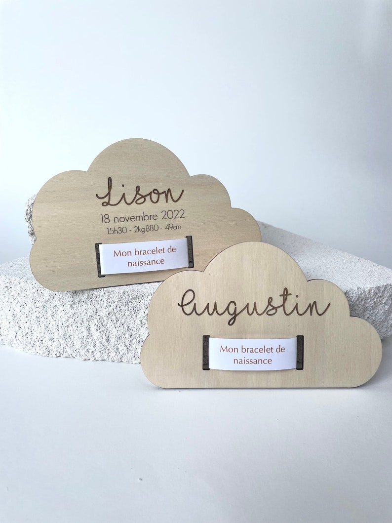 Puede incluir: Dos placas de madera en forma de nube con nombres, fechas de nacimiento y medidas grabadas. La placa superior dice "Lison 18 novembre 2022 15h30 - 2kg880 - 49cm" y la placa inferior dice "Augustin". Ambas placas tienen una peque&ntilde;a etiqueta blanca que dice "Mon bracelet de naissance".