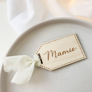 May include: A wooden place card with a stitched border and the name "Mamie" engraved in cursive. The card is tied with a light beige ribbon and is sitting on a light gray plate.