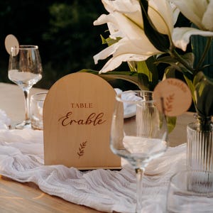 Op de afbeelding: Een houten tafelschild met het woord "Erable" in elegant schrift en het woord "TABLE" erboven. Onder het woord staat een kleine tak met bladeren. Het bord staat op een tafel met witte stof, glaswerk en witte bloemen.