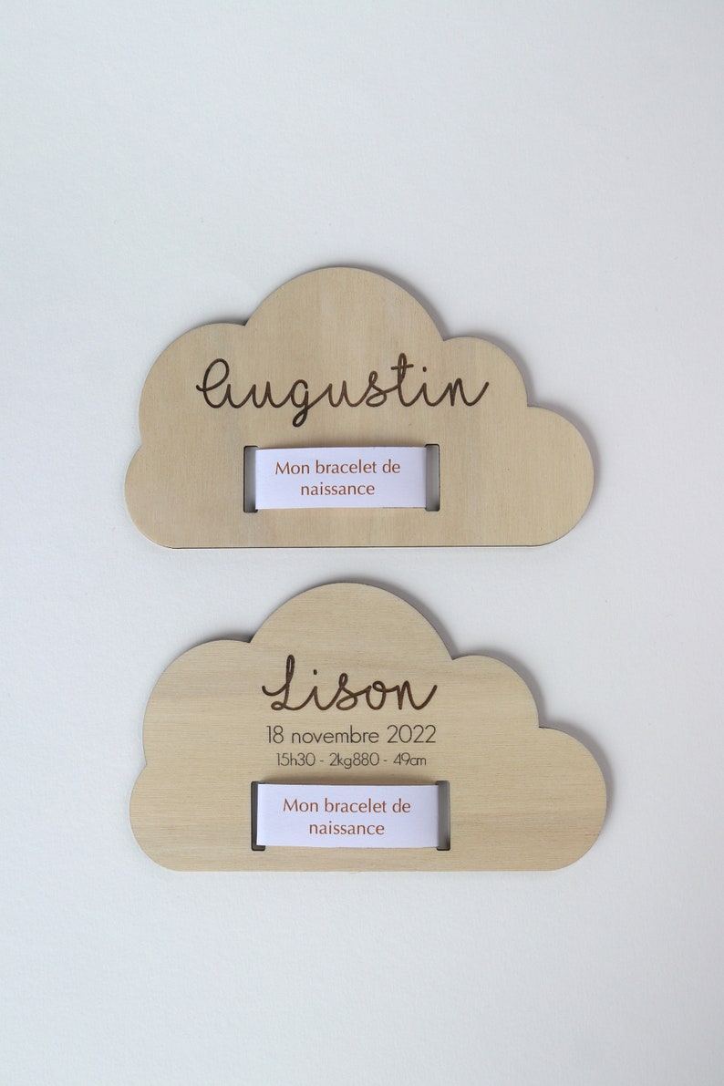 Puede incluir: Dos placas de madera en forma de nube con nombres, fechas de nacimiento y medidas grabadas. La placa superior dice "Augustin" y la placa inferior dice "Lison 18 de noviembre de 2022 15h30 - 2kg880 - 49cm". Ambas placas tienen una ranura para un brazalete.