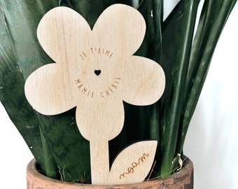 May include: A wooden flower-shaped plant marker with the words "JE T'AIME MAMIE CHERIE" and a heart engraved on the flower's center. The stem has the name "noam" engraved. The marker is placed in a terracotta pot with a green plant.
