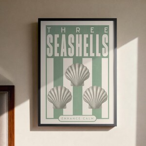 May include: A framed print with the words "THREE SEASHELLS" and "ENHANCE CALM". The artwork features three seashell illustrations on a striped background. The print is in a black frame, above a wooden bedside table.