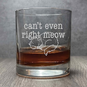 Puede incluir: Un vaso de vidrio transparente con el texto "can't even right meow" y una pequeña ilustración de un gato durmiendo. El vaso está lleno de un líquido ámbar oscuro.