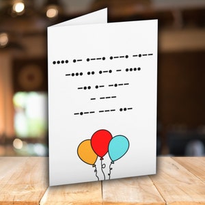 May include: White greeting card featuring a black Morse code message and three colourful balloons. The balloons are yellow, red, and blue, with black string. The card is on a wooden table with a blurred background.