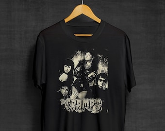 May include: Black t-shirt featuring a grayscale image of the band The Cramps. The graphic includes the band members and the band's name in a distressed font. The shirt is hanging on a wooden hanger.
