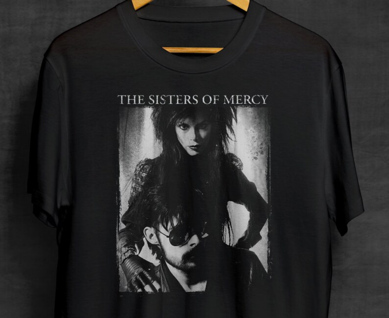May include: Black and white photo of The Sisters of Mercy band members on a black t-shirt. The band's name is printed in white above the photo.