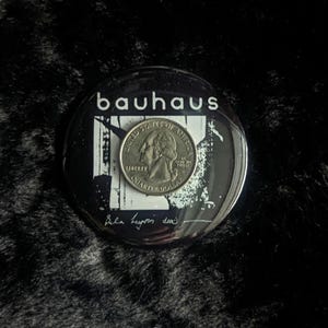 Op de afbeelding: Een zwart-witte knop met het woord "Bauhaus" in witte letters. De knop heeft een zwart-witte afbeelding van een gebouw met een kwartje erop. De tekst "Peter Murphy, 1982" is in zwarte inkt op de knop geschreven.
