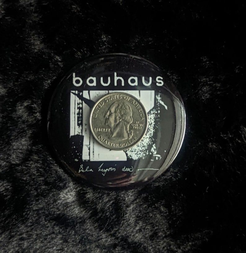 Op de afbeelding: Een zwart-witte knop met het woord "Bauhaus" in witte letters. De knop heeft een zwart-witte afbeelding van een gebouw met een kwartje erop. De tekst "Peter Murphy, 1982" is in zwarte inkt op de knop geschreven.