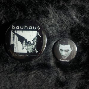 Puede incluir: Dos pines de bot&oacute;n en blanco y negro. El pin m&aacute;s grande presenta un murci&eacute;lago con el texto "Bauhaus" y " Bela Lugosi Dead". El pin m&aacute;s peque&ntilde;o presenta un retrato en blanco y negro de una persona con el texto "Damned".