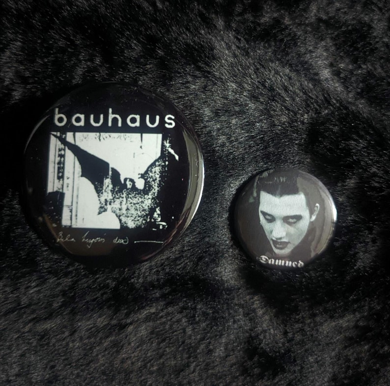 Puede incluir: Dos pines de bot&oacute;n en blanco y negro. El pin m&aacute;s grande presenta un murci&eacute;lago con el texto "Bauhaus" y " Bela Lugosi Dead". El pin m&aacute;s peque&ntilde;o presenta un retrato en blanco y negro de una persona con el texto "Damned".