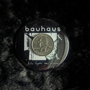 Puede incluir: Un bot&oacute;n blanco y negro con la palabra "bauhaus" en letras blancas. El bot&oacute;n tiene una imagen en blanco y negro de un edificio con una moneda de 25 centavos encima. La moneda de 25 centavos tiene las palabras "United States of America" y "Liberty" en ella. El bot&oacute;n tambi&eacute;n tiene las palabras "Bella begins down" escritas con tinta negra.