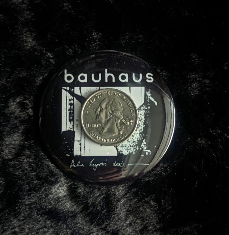 Puede incluir: Un bot&oacute;n blanco y negro con la palabra "bauhaus" en letras blancas. El bot&oacute;n tiene una imagen en blanco y negro de un edificio con una moneda de 25 centavos encima. La moneda de 25 centavos tiene las palabras "United States of America" y "Liberty" en ella. El bot&oacute;n tambi&eacute;n tiene las palabras "Bella begins down" escritas con tinta negra.