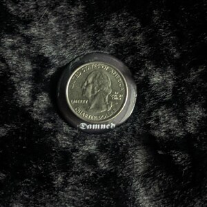 Puede incluir: Un bot&oacute;n negro con una moneda de cuarto de d&oacute;lar estadounidense de plata incrustada en el centro. La moneda presenta el perfil de George Washington y las palabras "United States of America", "Liberty" y "Quarter Dollar". El bot&oacute;n tiene la palabra "Damned" impresa en blanco debajo de la moneda.