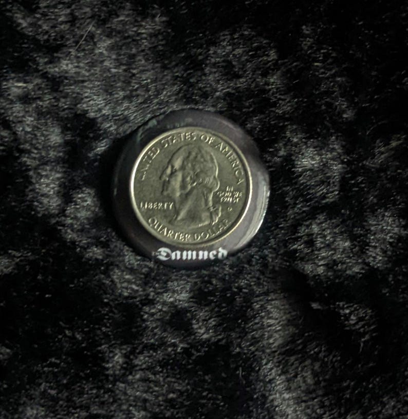 Puede incluir: Un bot&oacute;n negro con una moneda de cuarto de d&oacute;lar estadounidense de plata incrustada en el centro. La moneda presenta el perfil de George Washington y las palabras "United States of America", "Liberty" y "Quarter Dollar". El bot&oacute;n tiene la palabra "Damned" impresa en blanco debajo de la moneda.