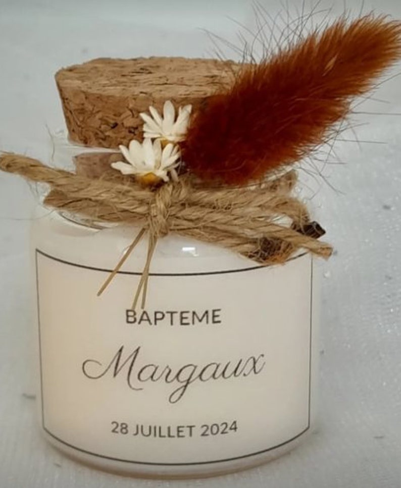 May include: A small glass jar with a cork lid, decorated with dried flowers and twine. The jar has a white label with the text "BAPTEME Margaux 28 JUILLET 2024".