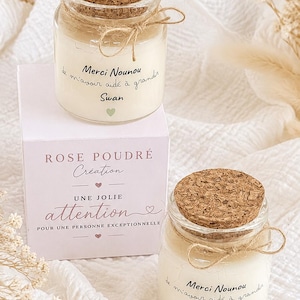 Peut inclure: Deux pots en verre avec couvercles en liège contenant de la cire blanche, chacun noué avec de la ficelle. Les étiquettes portent l'inscription "Merci Nounou de m'avoir aidé à grandir" avec le nom "Swan" et un cœur vert. Une boîte blanche indique "ROSE POUDRÉ Création" et "UNE JOLIE attention".