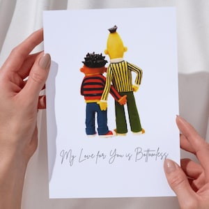 Op de afbeelding: Een witte kaart met een cartoon illustratie van twee figuren die elkaars hand vasthouden. Eén is geel met een gestreept shirt, de ander is oranje en zwart. De tekst op de kaart is "My Love for You is Bottomless."