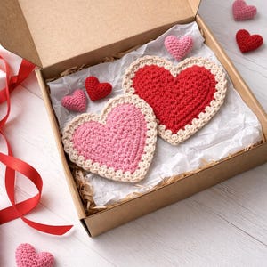 May include: A cardboard box contains two large crocheted hearts, one red and one pink, with cream-colored borders. Several smaller crocheted hearts in red and pink are scattered around the box. The hearts are presented on white paper and wood shavings.