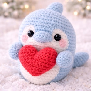 Puede incluir: Un peluche de delfín tejido a mano en azul claro, blanco y rojo. El delfín sostiene un corazón rojo, con ojos negros y mejillas rosadas. El fondo es blanco suave y borroso con luces bokeh, sugiriendo un tema de amor.