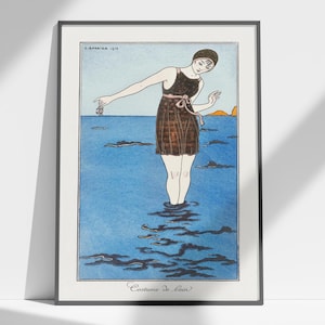 May include: Framed vintage art print featuring a woman in a brown swimsuit standing in blue water. The artwork, titled "Costume de bain," includes text and is set against a light blue sky with a small island in the distance. The artist's name and the year 1911 are also visible.