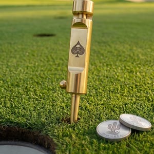 May include: A cylindrical brass golf divot tool featuring a spade and skull engraving, standing upright in the grass. Two circular silver metal ball markers with a cactus logo and the text Made in USA are positioned next to the tool.
