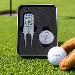 May include: A golf gift set featuring a divot tool, ball marker, and a Titleist golf ball. The divot tool and ball marker are in a metal case. The divot tool is engraved with "K.C.C. 2026". The golf ball is white with the number "1".