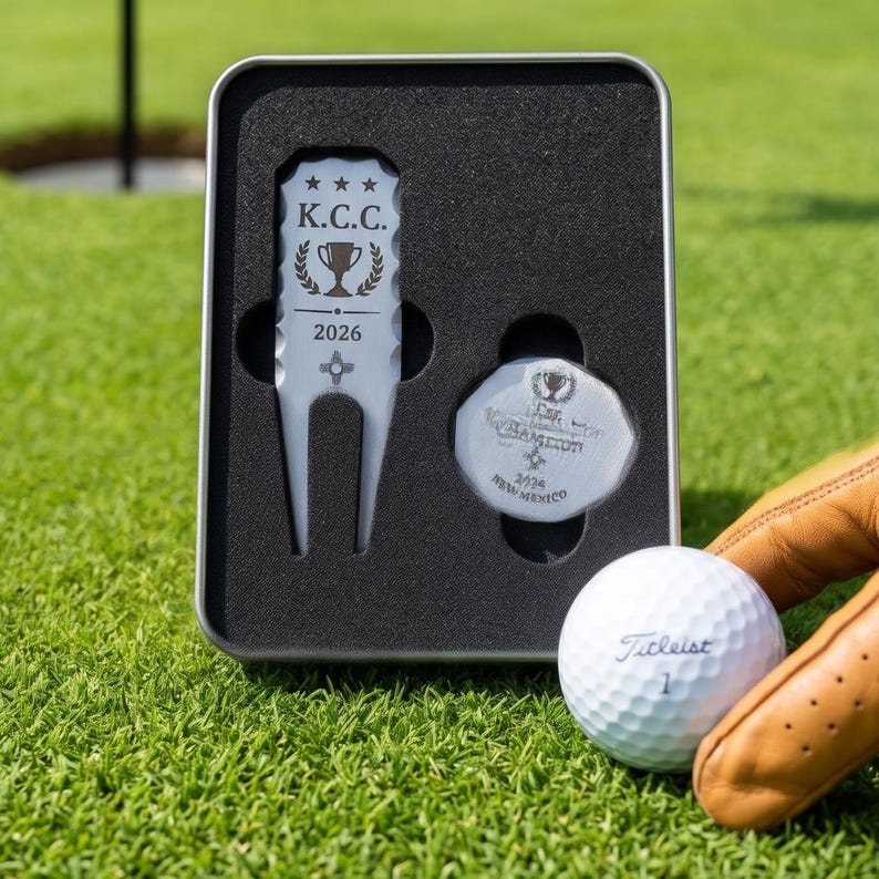 May include: A golf gift set featuring a divot tool, ball marker, and a Titleist golf ball. The divot tool and ball marker are in a metal case. The divot tool is engraved with "K.C.C. 2026". The golf ball is white with the number "1".
