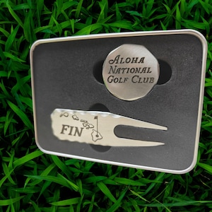 May include: A silver golf ball marker and divot repair tool set inside a black metal tin. The marker is round with "Aloha National Golf Club" text. The tool has "FIN" and a Hawaiian island graphic.