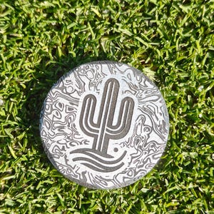 May include: A round, silver-colored golf ball marker with a desert cactus design. The marker has a textured pattern and is set against a background of green grass.