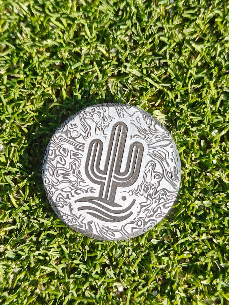 May include: A round, silver-colored golf ball marker with a desert cactus design. The marker has a textured pattern and is set against a background of green grass.
