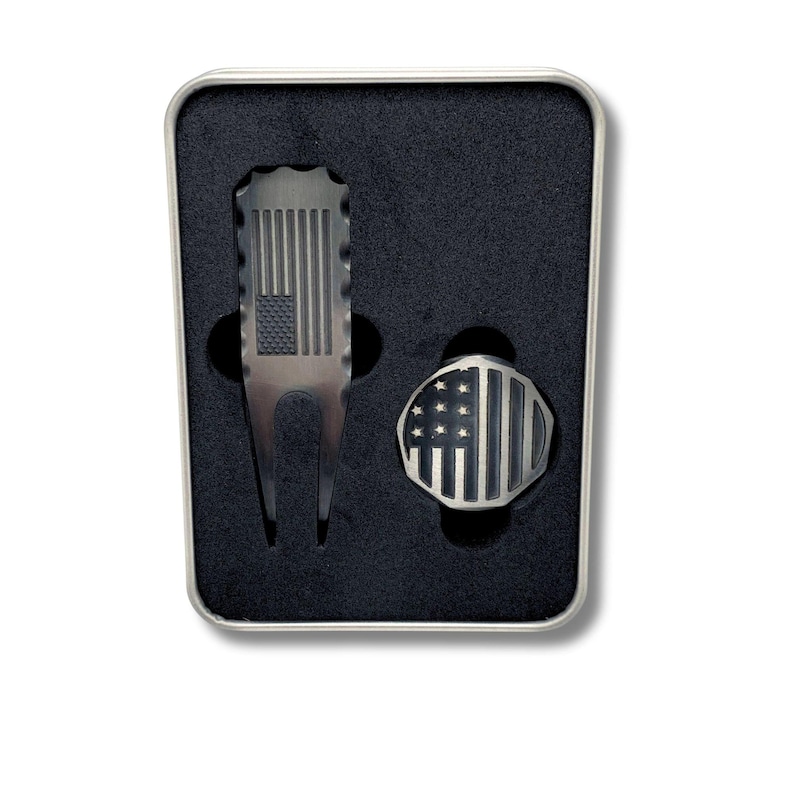 Close-up of a custom engraved golf divot tool and a ball marker, each with an American flag design, in a black foam-lined metal gift box.