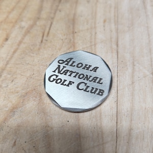 Close-up of a silver-toned, octagonal golf ball marker that reads "ALOHA NATIONAL Golf Club" in black text.