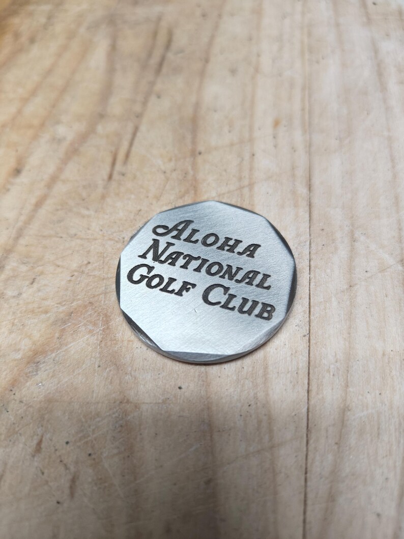Close-up of a silver-toned, octagonal golf ball marker that reads "ALOHA NATIONAL Golf Club" in black text.