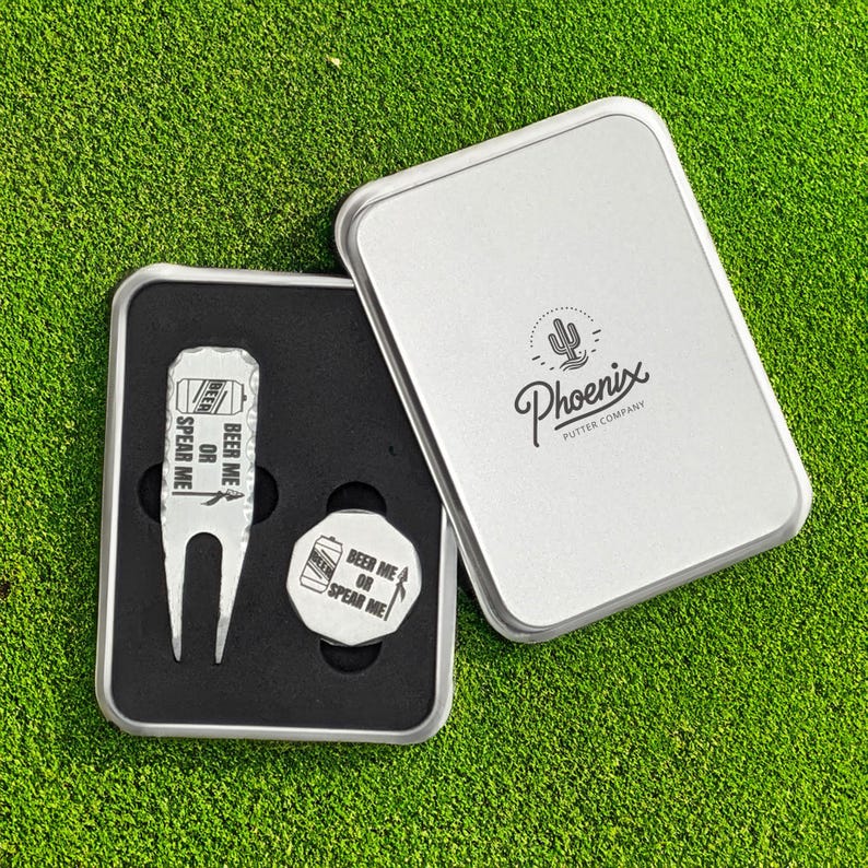 May include: A silver golf divot repair tool and ball marker set in a metal case. The tool and marker have the text "BEER ME OR SPEAR ME!". The case is silver with the Phoenix Putter Company logo.