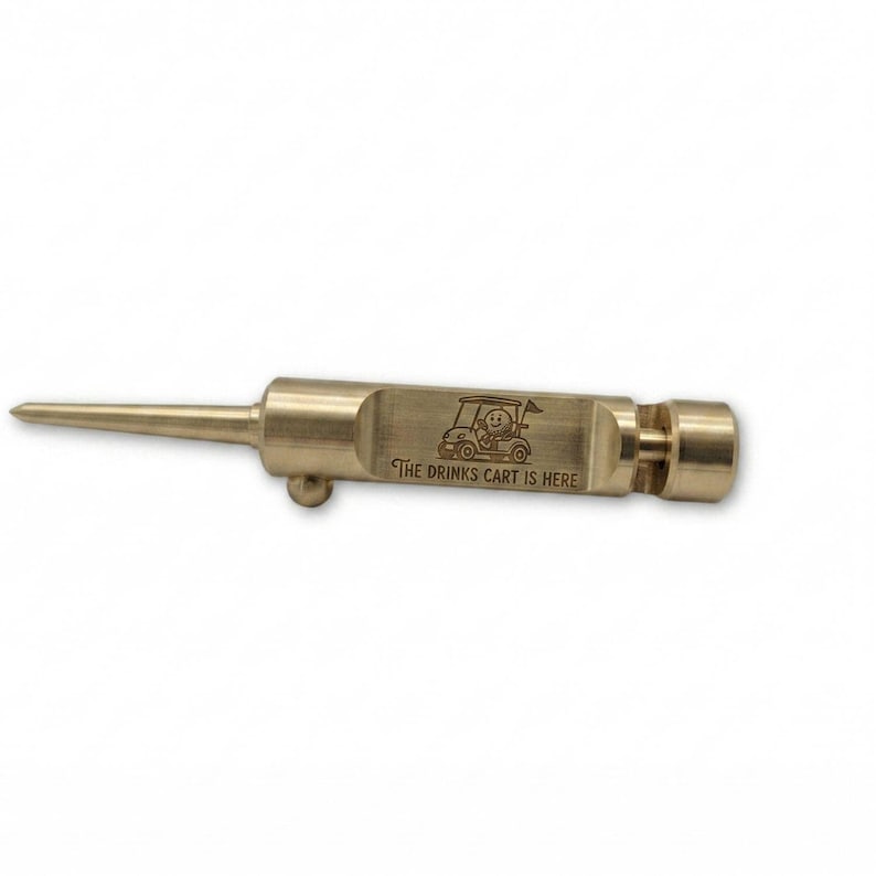Close-up of a brass, retractable golf divot tool on a light wooden table. A brown book is blurred in the background.