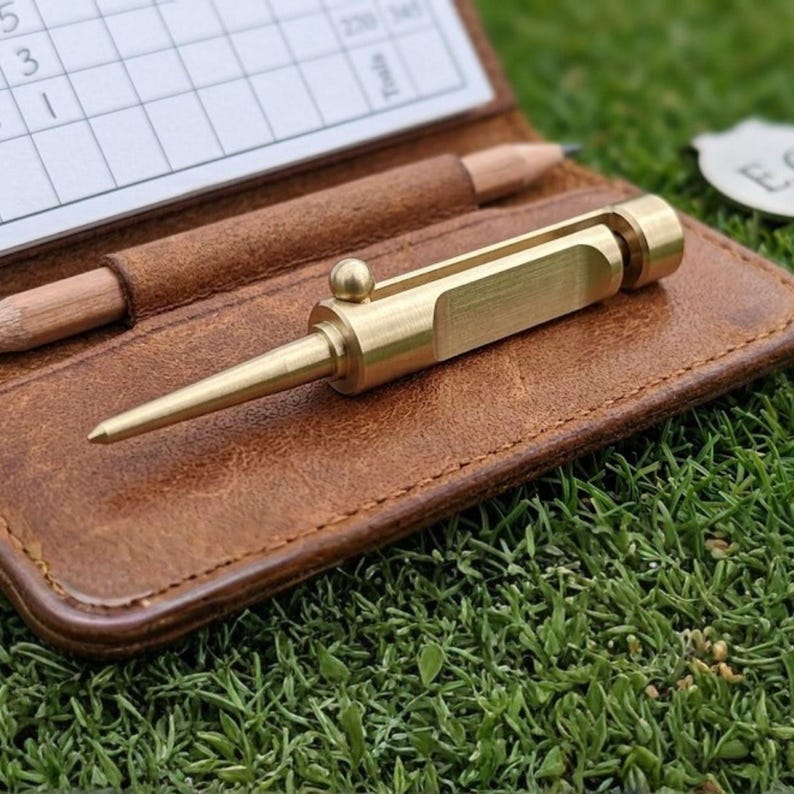 Close-up of a gold brass divot tool on a brown leather scorecard holder with a pencil, resting on green grass.