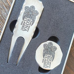 Close-up of a custom engraved golf divot tool and a matching ball marker resting in dark gray foam packaging.