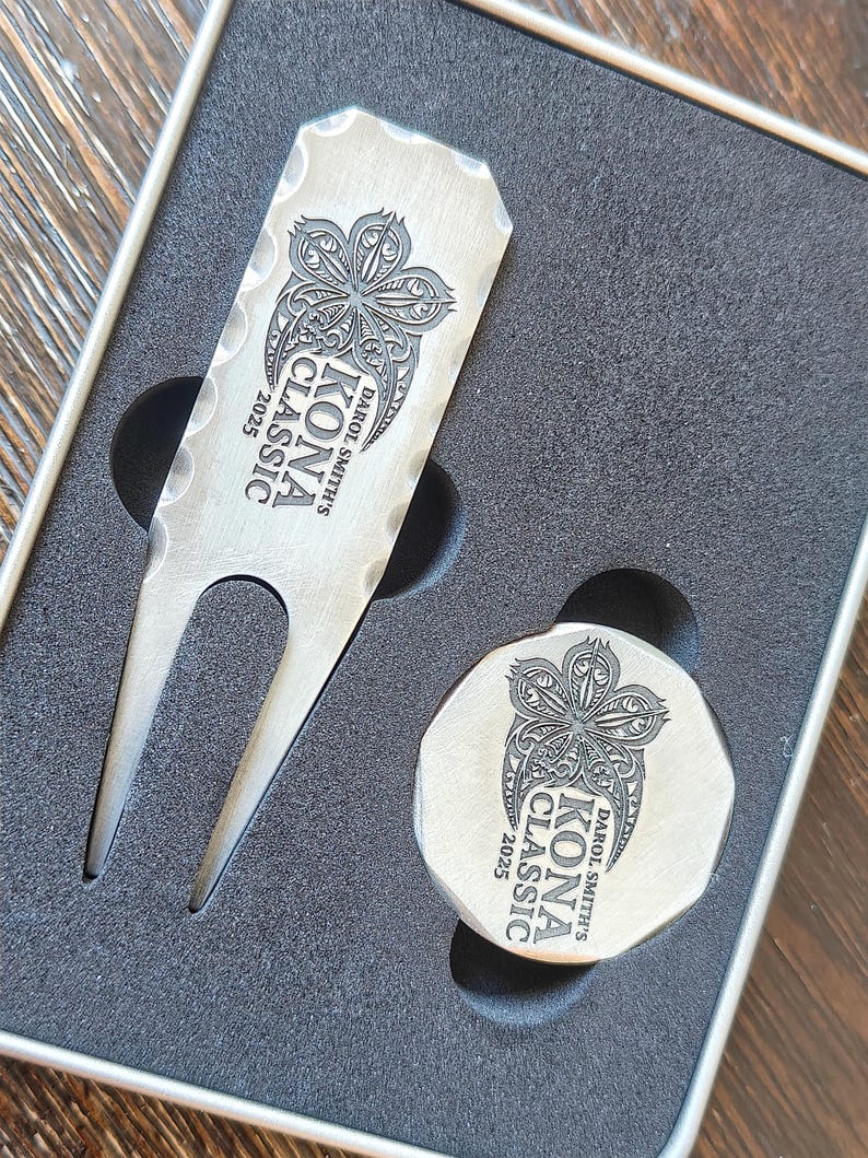 Close-up of a custom engraved golf divot tool and a matching ball marker resting in dark gray foam packaging.