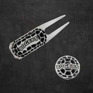 A silver engraved golf divot tool and ball marker with the word "BOGEYS" in black with a cracked pattern.