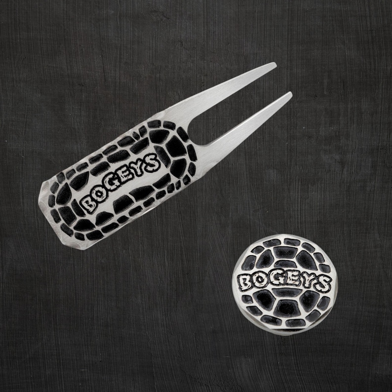 A silver engraved golf divot tool and ball marker with the word "BOGEYS" in black with a cracked pattern.